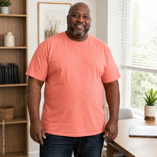 A cheerful man wearing a coral t-shirt stands in a bright, modern home office, radiating positivity and confidence.