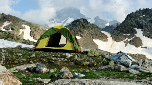 A yellow-green tent pitched on a rocky alpine ridge surrounded by snow patches, wildflowers and towering peaks, conveying high-altitude adventure, solitude and remote outdoor wilderness camping.