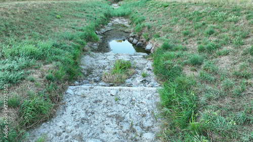 River drought dried stream dry shallow waterway Trusovicky creek climate change bed with cracked mud, exposed rocks, sediment, tall grasses, reeds, shrubs, and trees clear, sunny, cracked soil
