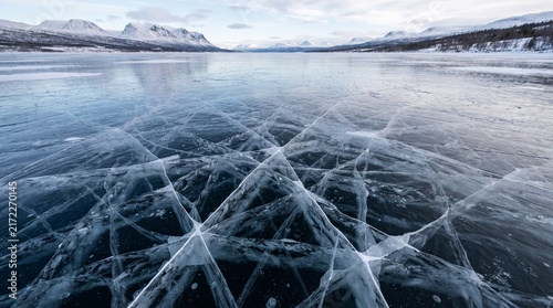 Stunning wide-angle shot of a vast, frozen lake in a Nordic landscape with complex, intricate crack patterns spreading across the dark, clear ice leading toward distant snowy mountains.