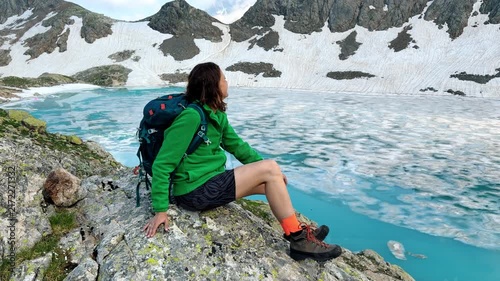 Backpacker Hiker Sitting on Rocky Ledge Over Turquoise Glacier Lake in Alpine Mountains and Enjoys The View