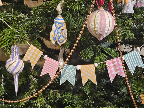 Festive banner hangs on green pine branches. Colorful ornaments dangle beside the banner. Gold beads trail along the branches. Soft pastel patterns decorate each banner section