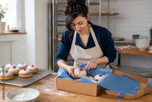 Baker Packs Fresh Doughnuts: A smiling baker, wearing an apron, carefully places freshly made doughnuts into a cardboard box, ready for delivery.