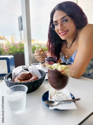 Young brunette woman with glasses sits at an outdoor cafe table, smiling at the camera while eating authentic Sicilian granita with a fresh brioche pastry served in a basket for breakfast or brunch.