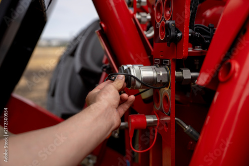 Male hand removing protective rubber dust cap from hydraulic quick release coupling valve on modern red agricultural tractor in field.