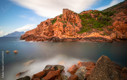Long-exposure view of the Genoese Tower of Porto above red granite cliffs and calm Mediterranean water on Corsica, France.