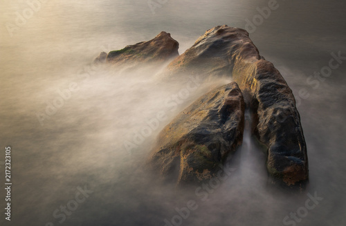 Long-exposure detail of coastal rocks surrounded by silky Mediterranean water on the rugged shoreline of Porto, Corsica, France.