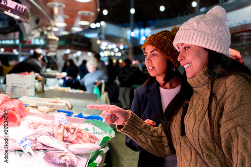 Obraz na płótnie Two smiling women wearing knit hats and warm sweaters pointing at fresh fish and