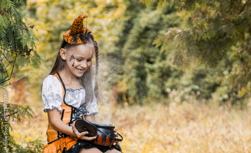 Fotografie Girl in a Costume Sits in a Field During Autumn, Surrounded by Colorful Trees an