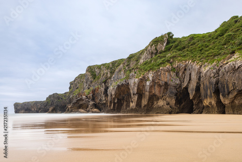 Playa de Andrín beach showing golden sand and jagged limestone cliffs with sea caves covered in lush greenery. Llanes, Costa Verde, Asturias, Northern Spain.