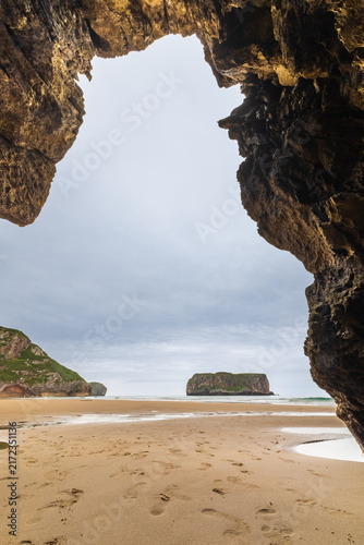 Looking out from inside a coastal cave toward the golden sand of Playa de Andrín, the ocean, and a rocky island on a moody day. Costa Verde, Asturias, Northern Spain.