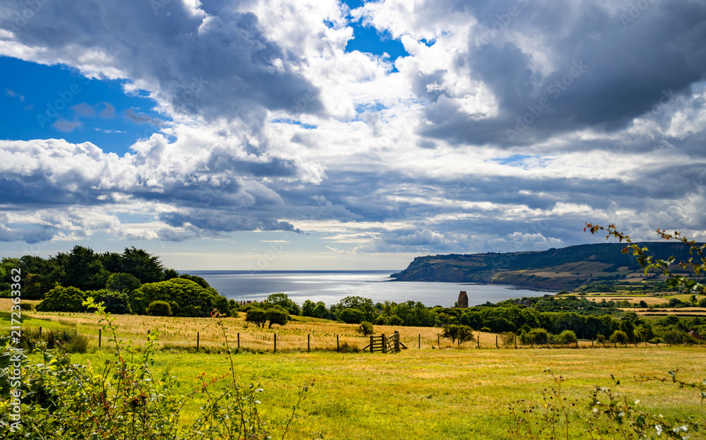 Obraz premium Dramatic clouds and sunlight over fields and the North Sea near Robin Hood’s Bay, with the rugged Yorkshire coastline stretching towards Ravenscar, North Yorkshire, England.