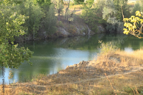 Scenic landscape of a rocky quarry lake with clear water, surrounded by trees and dry grass on a sunny summer day