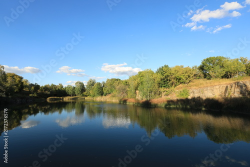 Scenic landscape of a rocky quarry lake with clear water, surrounded by trees and dry grass on a sunny summer day