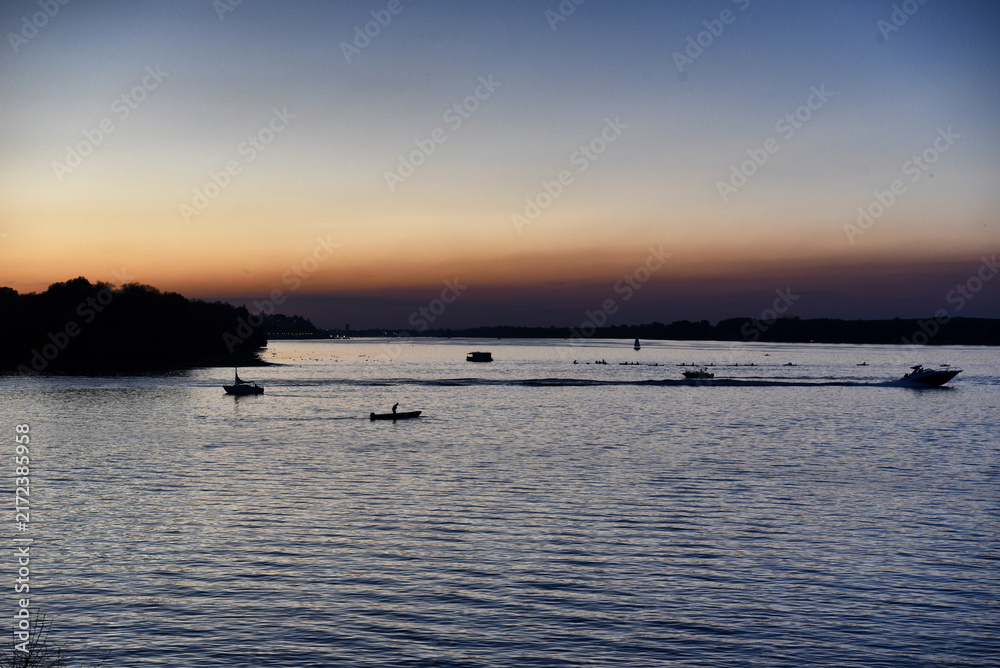 Obraz premium Boats sailing on a wide river during twilight dusk with orange and blue sky horizon
