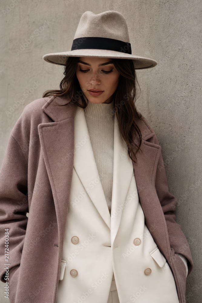 Fototapeta premium Elegant woman in chic beige fedora and oversized camel coat posing confidently against neutral urban wall, stylish autumn fashion with white double-breasted blazer and ribbed turtleneck, moody
