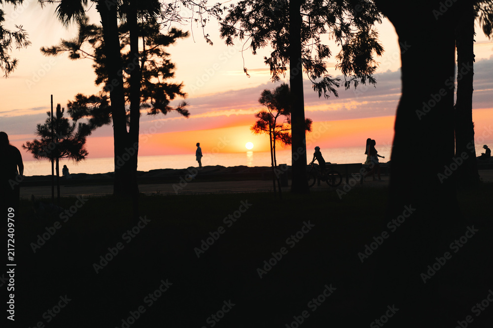 Naklejka premium Sunset silhouette of people outdoors along a beach by water, warm sky and calm horizon on soft sand and gentle breeze