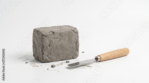 Gray clay block with sculpting tool on light surface near window, raw material and instrument symbolizing the beginning of creative artistry and hands‑on craftsmanship.