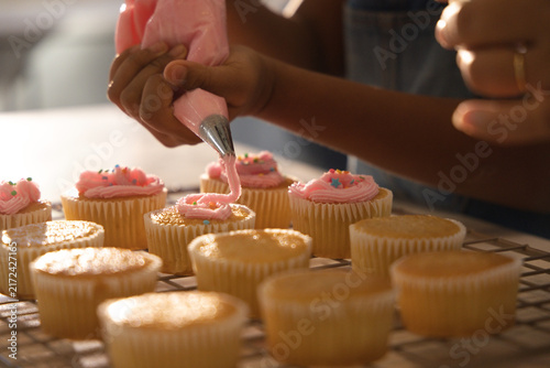 African American mom guiding youth in denim at home kitchen squeezing piping bag icing cupcakes