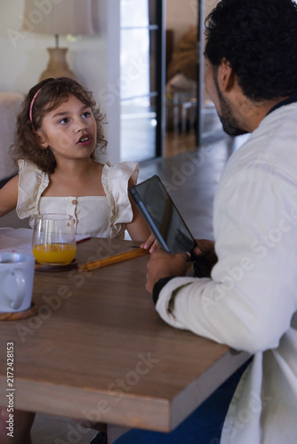 African American dad showing tablet, child in ruffled dress sipping OJ at wooden table, copy space