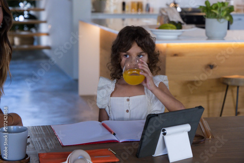African American schoolgirl drinking orange juice and doing homework at kitchen table with tablet