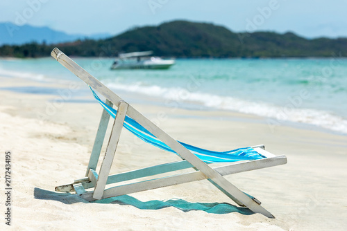 Foto Wooden beach lounge chair with blue fabric rests on the sandy shore, with gentle ocean waves lapping nearby and a boat visible in the backdrop of hills