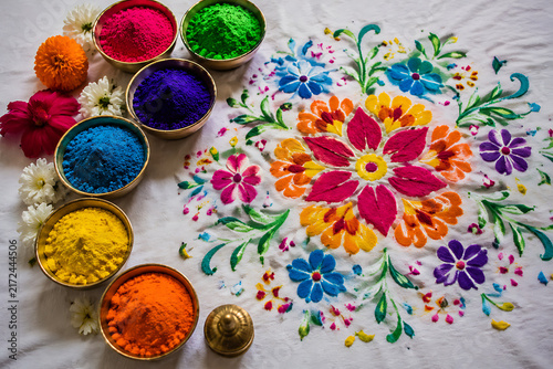 Intricate Floral Rangoli Pattern Design Surrounded by Bowls of Bright Holi Gulal Powders and Fresh Flowers on White Fabric