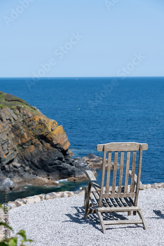 A wooden deckchair on a gravel garden patio overlook the coast, showing blue sea, cloudless sky, and a rocky coastline.