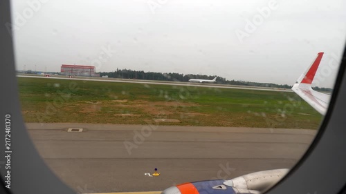 Airplane taxiing on airport runway viewed through raindrops on passenger window on overcast rainy day with another plane in background