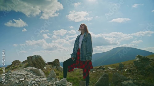 Girl stands on rocky ground with mountains in the background on a sunny day with clouds in the sky