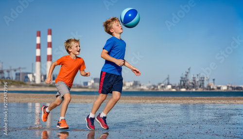 Two children play with a colorful ball in the foreground while blurred industrial smokestacks and coastal buildings rise against a bright blue sky.