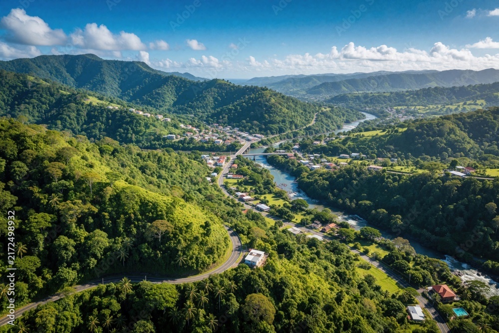 Fototapeta premium Aerial view of rich green mountains in Puerto Rico. Small town sits near winding river in tropical valley. Winding road leads through dense jungle, rural landscape. Scenic landscape shows mountain