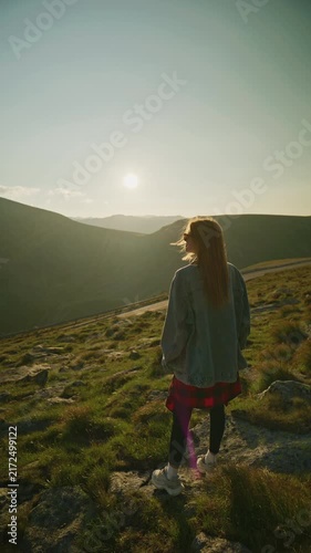 Woman stands on a hillside and looks at the sunset behind mountains during evening hours in a natural setting