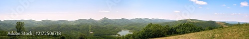 Ultra-wide panorama of endless green mountains and hills. A lake and small buildings visible in the valley. Clear blue sky, summer landscape.