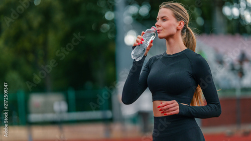 Fotografie Fit woman in black sportswear drinking water from bottle after workout on stadiu