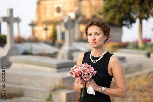 Canvastavla Sorrowful woman in mourning black dress standing in cemetery with flowers in han