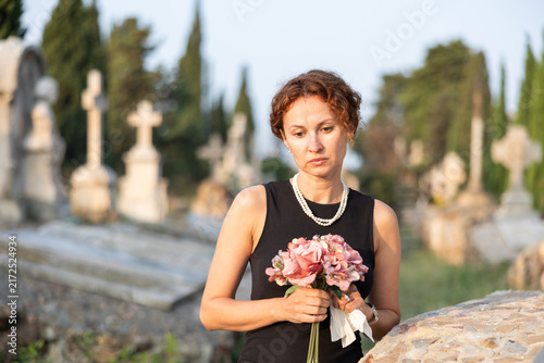 Foto Sorrowful woman in mourning black dress standing in cemetery with flowers in han