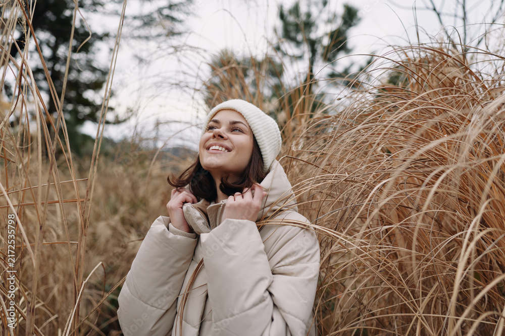 Fototapeta premium Woman in a warm coat and beanie gazes upward among tall dried grass in a winter meadow, outdoor portrait with soft natural light and a peaceful, hopeful mood.
