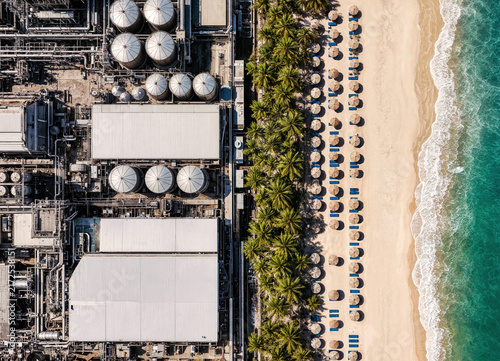 High-angle drone shot capturing the stark contrast between complex industrial metal tanks, a pristine sandy coastline, palm trees, and clear turquoise water.