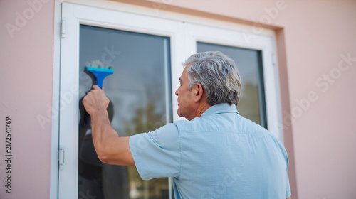 Middle-Aged Man Cleaning Glass Window with Squeegee at Home - Professional Household Maintenance and Chores