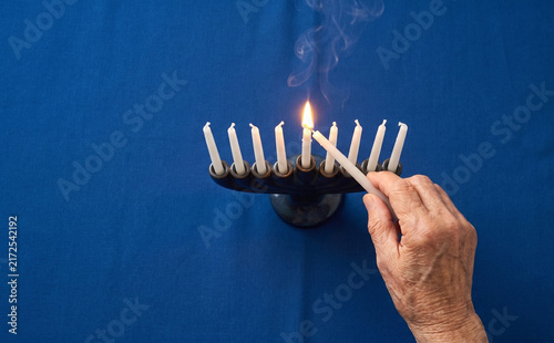 Obraz na płótnie Elderly hands lighting a menorah candle for Hanukkah celebration on blue background