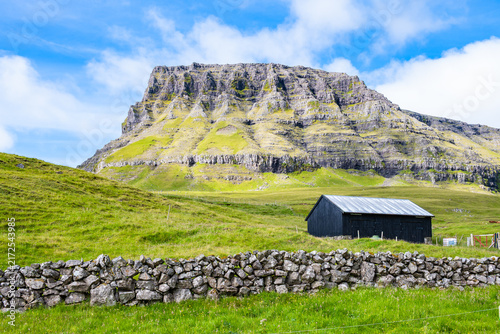 Black wooden farm building set against a dry stone wall and dramatic steep mountain cliffs near Gasadalur village, Faroe Islands, Denmark