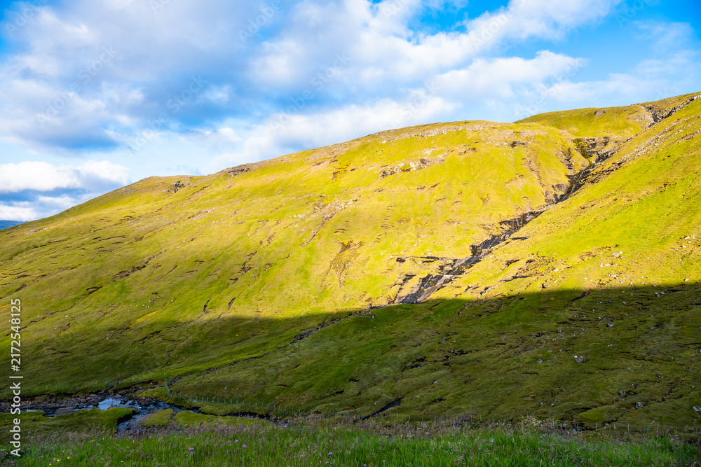 Fototapeta premium Grassy mountain slope with small streams flowing down, valley in shadow, blue sky with white clouds, Vagar, Faroe Islands, Denmark