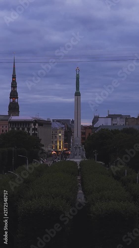 Aerial view gliding above tree lined alley toward Freedom Monument and Saint Peter Church tower in Riga, Latvia during evening twilight.