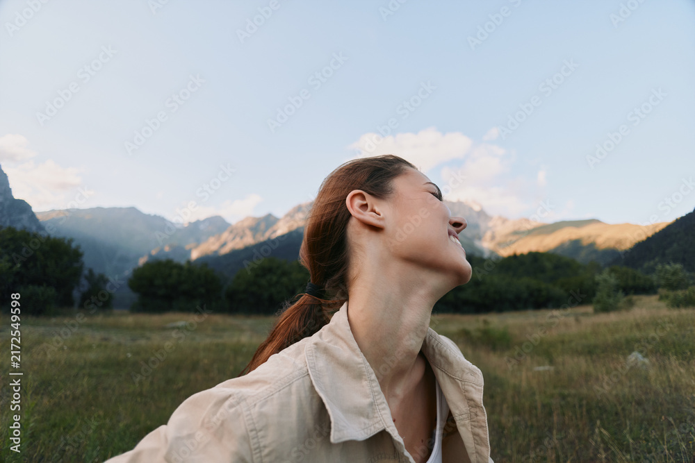 Fototapeta premium Happy woman outdoors in nature, smiling with eyes closed, enjoying fresh air, casual clothing, green meadow and mountains background, peaceful moment