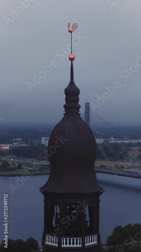 Aerial view of the historic Riga Cathedral spire with its weathercock above the Daugava River and cityscape at dusk in Riga, Latvia.