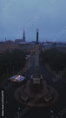 An aerial shot slowly circles the historic Freedom Monument and surrounding park in Riga, Latvia during a dark stormy evening.