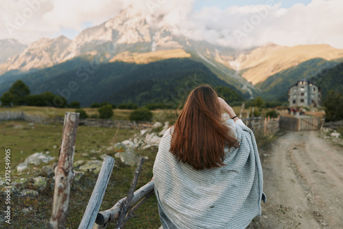 Bild auf Leinwand Young woman with long brunette hair walks along a mountain trail, wrapped in a gray shawl as distant peaks loom, a tranquil countryside scene suggesting adventure and contemplation