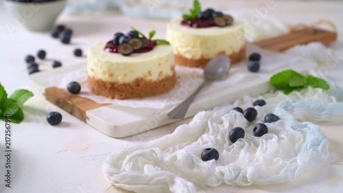 Two creamy mini cheesecakes sit on a rustic board, elegantly topped with vibrant red compote, fresh blueberries, and mint leaves under soft natural light highlighting their smooth texture.
