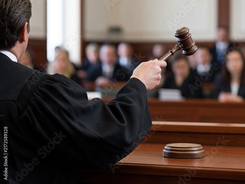 A judge in a black robe holds a wooden gavel in a courtroom with people sitting in the background.